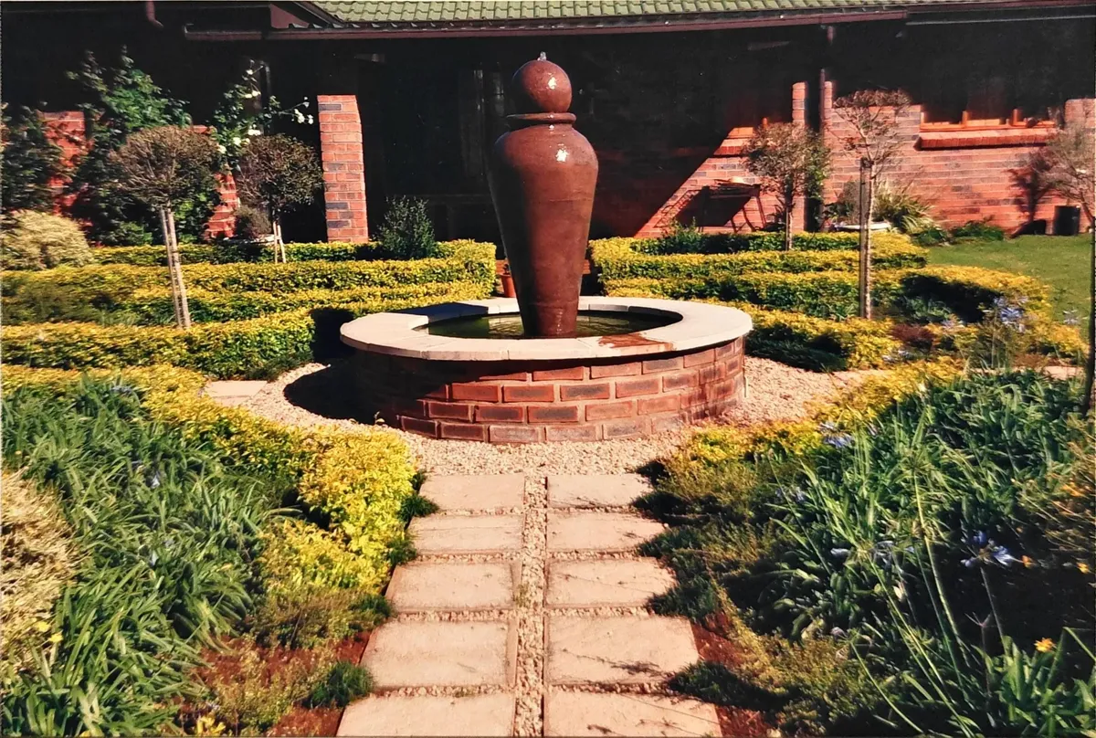 A formal garden design featuring a central circular stone water feature with a large brown urn fountain, flanked by golden hedges.